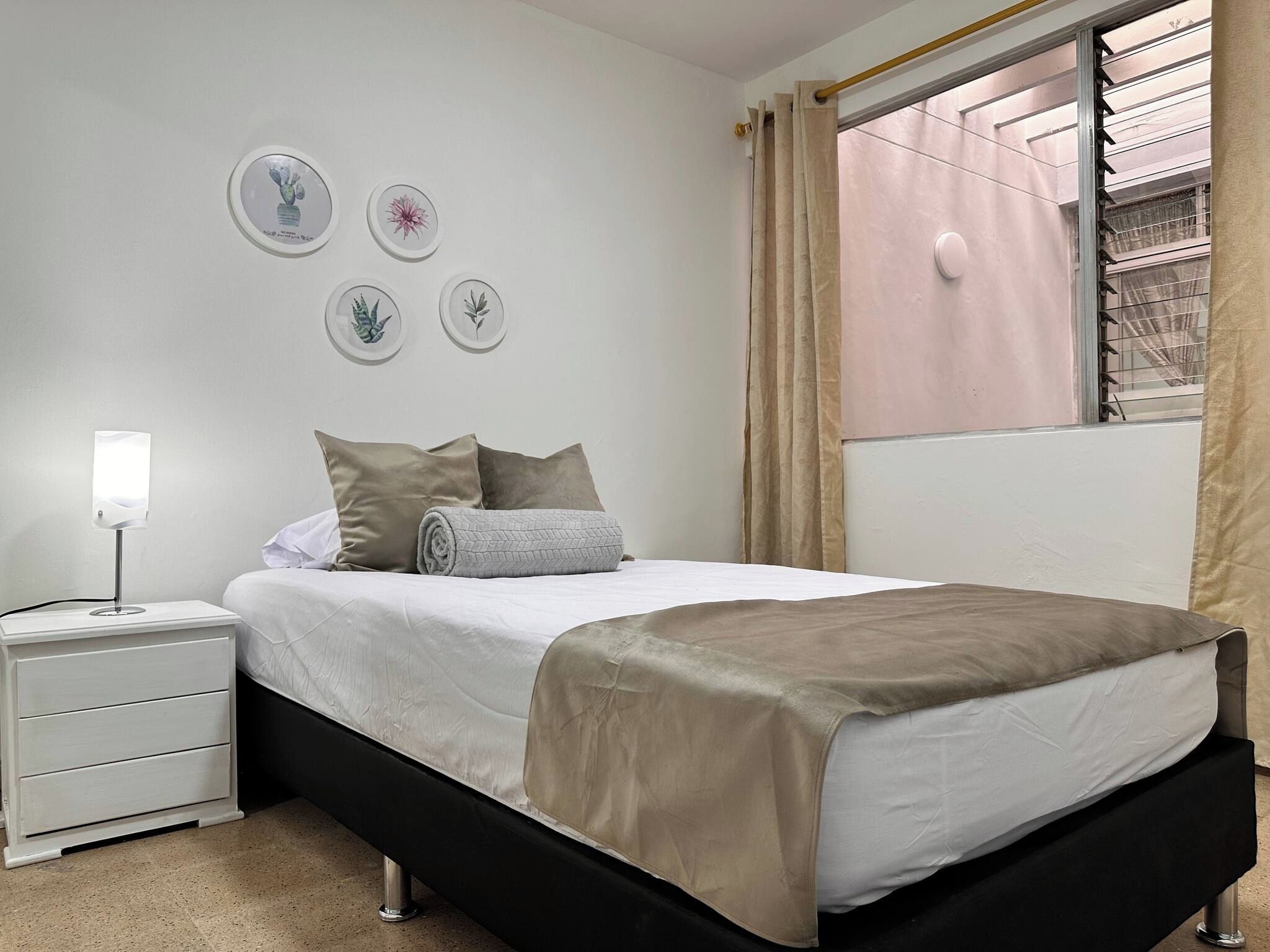 A neatly made bed with a white and beige bedspread, a bedside lamp, and a white nightstand with drawers.