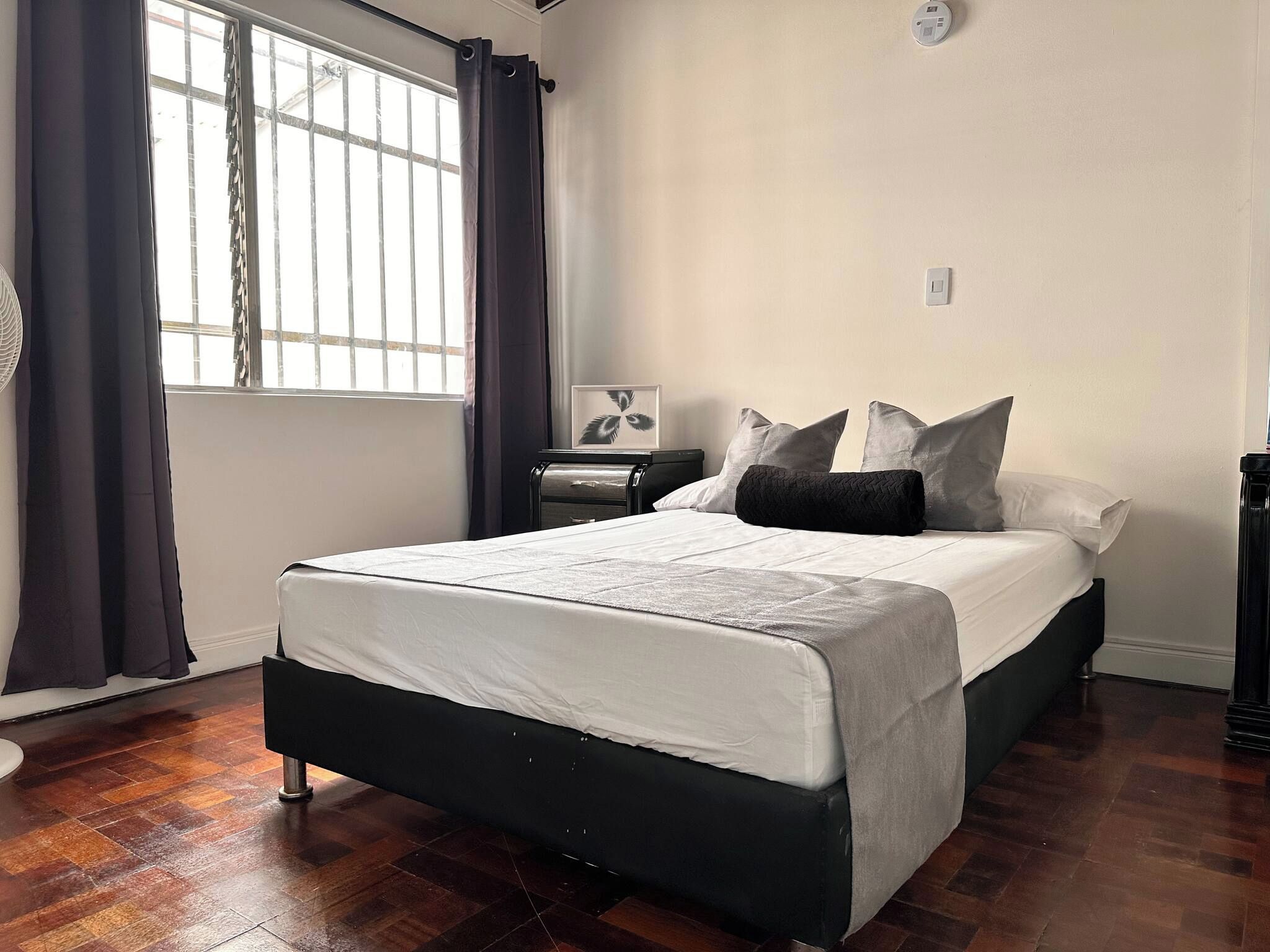 A bedroom with a bed, a nightstand, a wall clock, and a window with curtains.