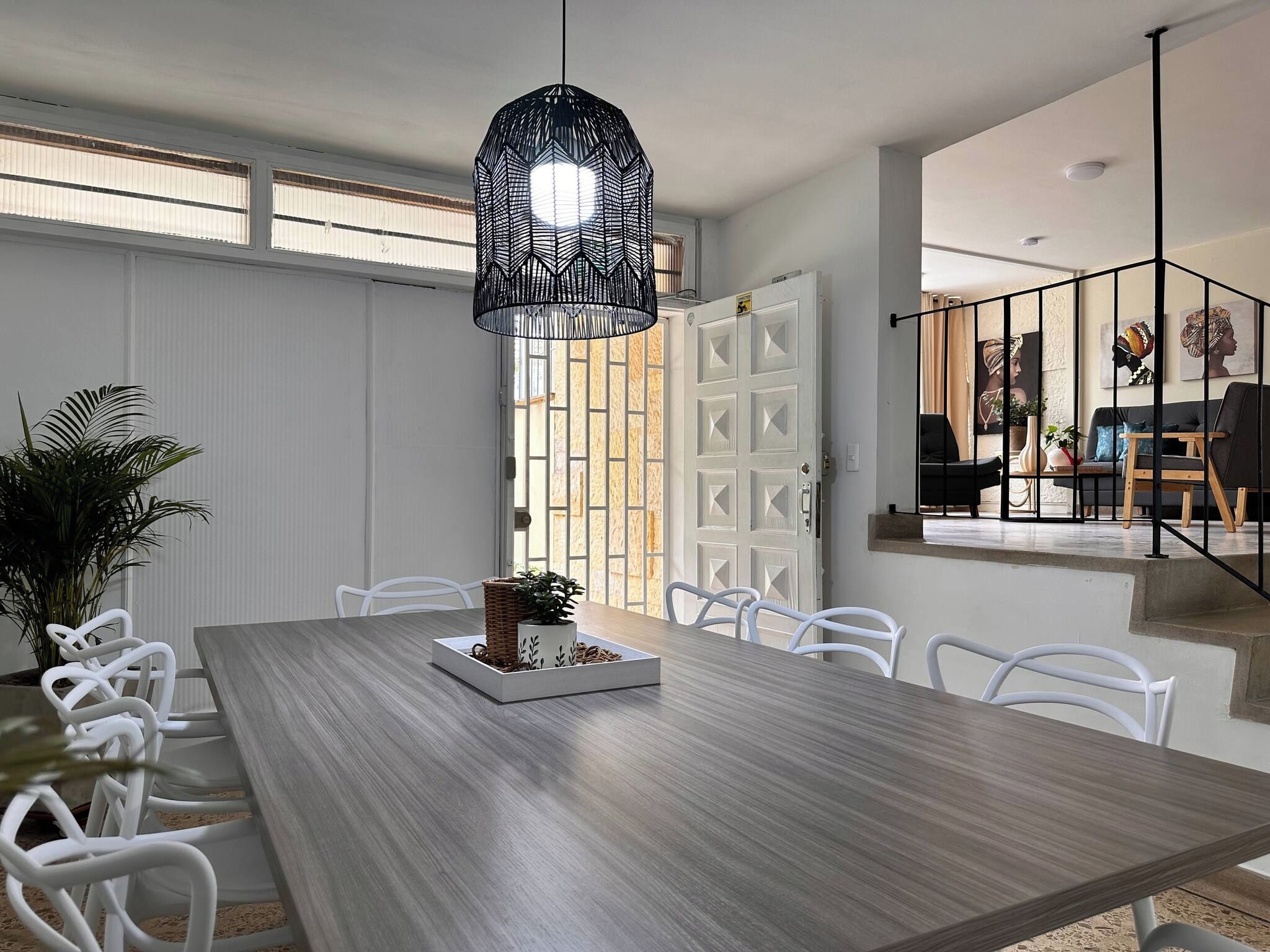 Un comedor con una larga mesa de madera, sillas blancas, una lámpara colgante negra y una planta en maceta.