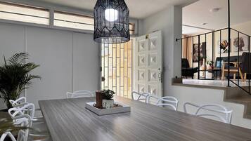 Un comedor con una larga mesa de madera, sillas blancas, una lámpara colgante negra y una planta en maceta.