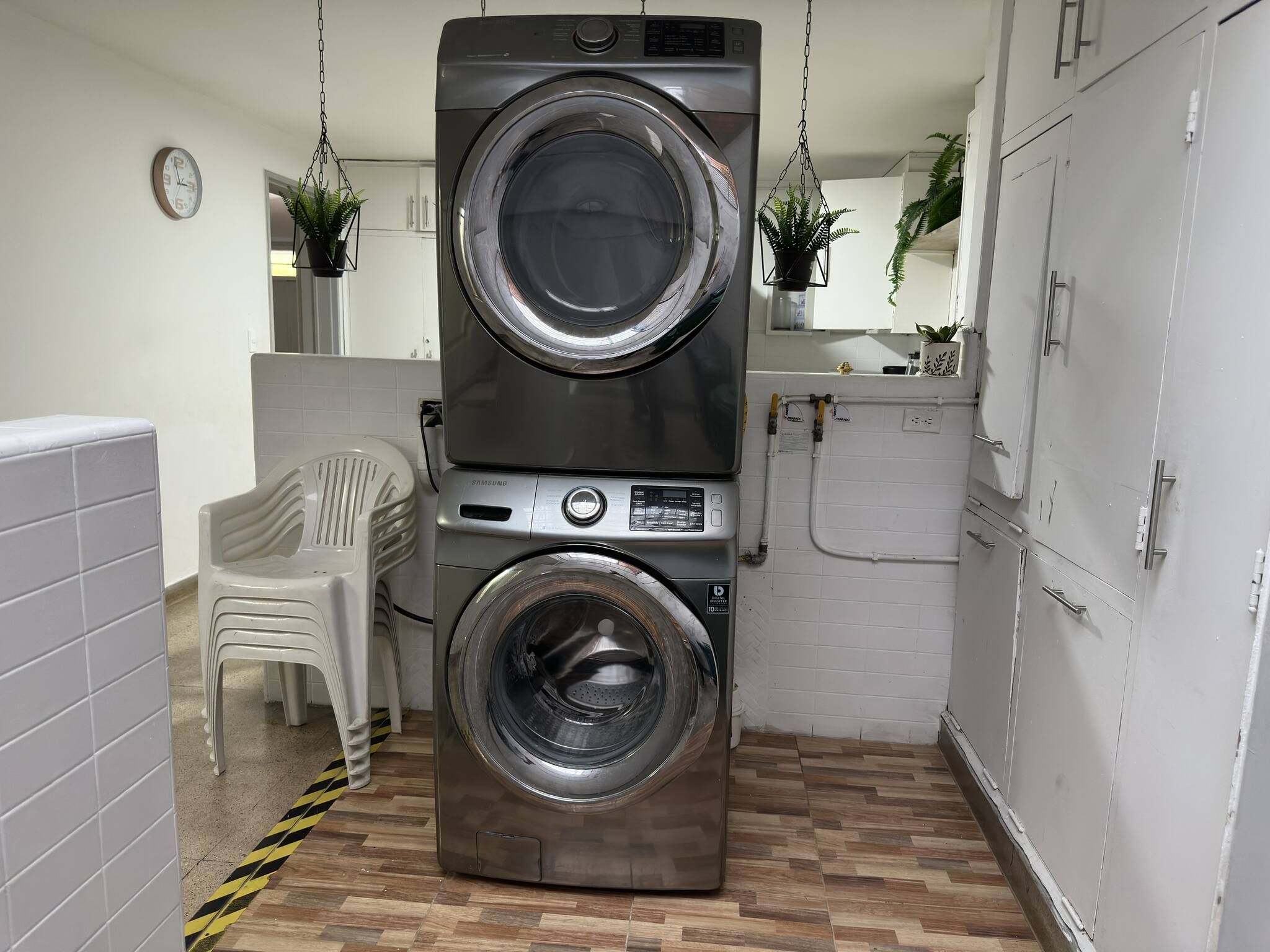 A compact laundry room with a stacked washer and dryer, a plastic chair, and a clock on the wall.