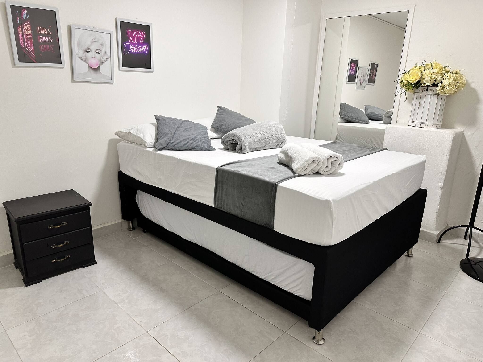 Un dormitorio moderno con una cama negra y blanca, almohadas grises, una mesita de noche pequeña y un espejo que refleja un jarrón con flores.