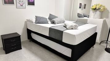 Un dormitorio moderno con una cama negra y blanca, almohadas grises, una mesita de noche pequeña y un espejo que refleja un jarrón con flores.