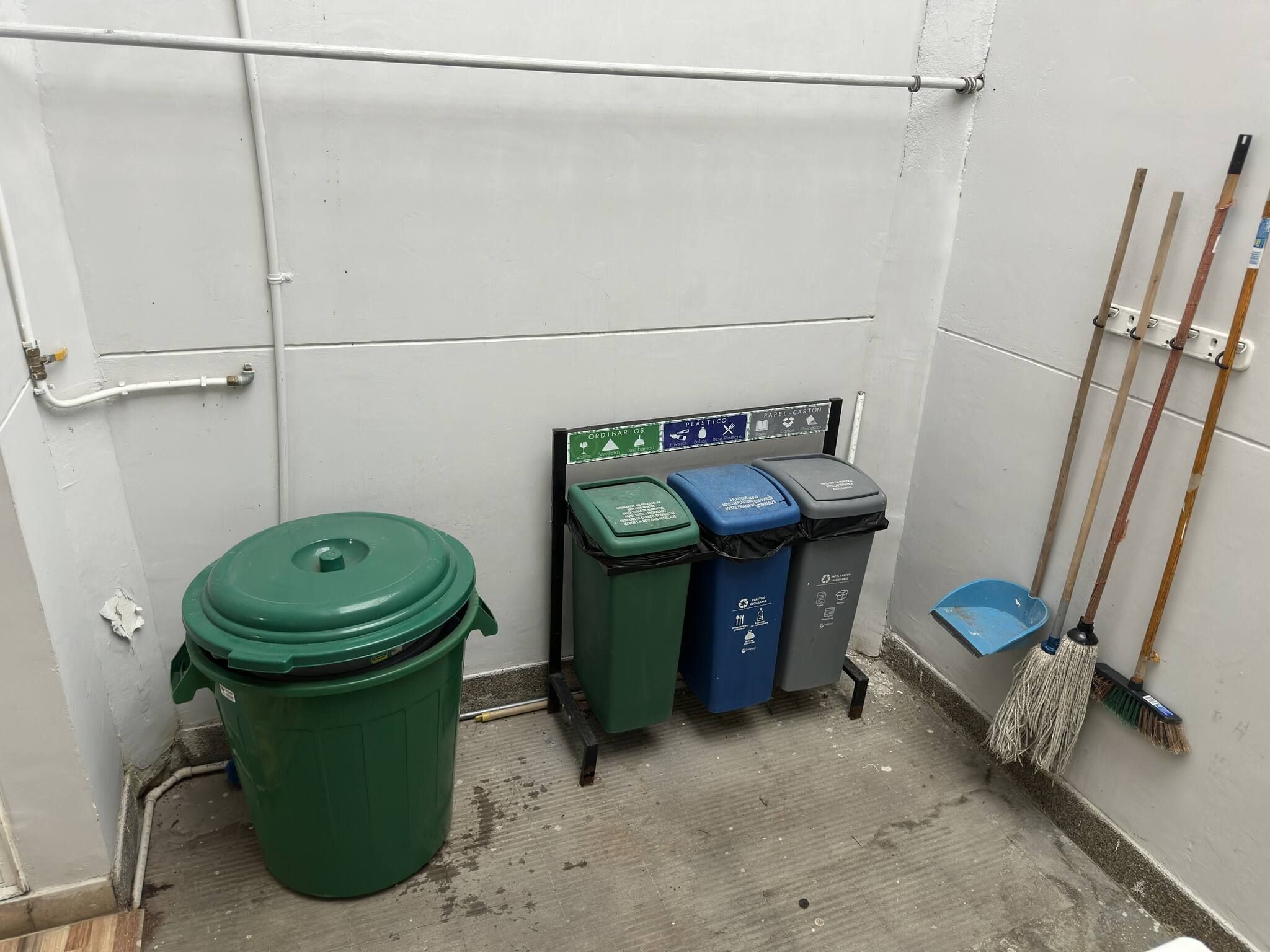 A small, enclosed utility room with a green trash bin, three recycling bins, and a broom leaning against the wall.