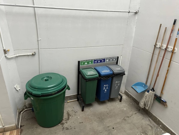 Un pequeño cuarto de servicio cerrado con un bote verde para basura, tres contenedores de reciclaje y una escoba apoyada en la pared.
