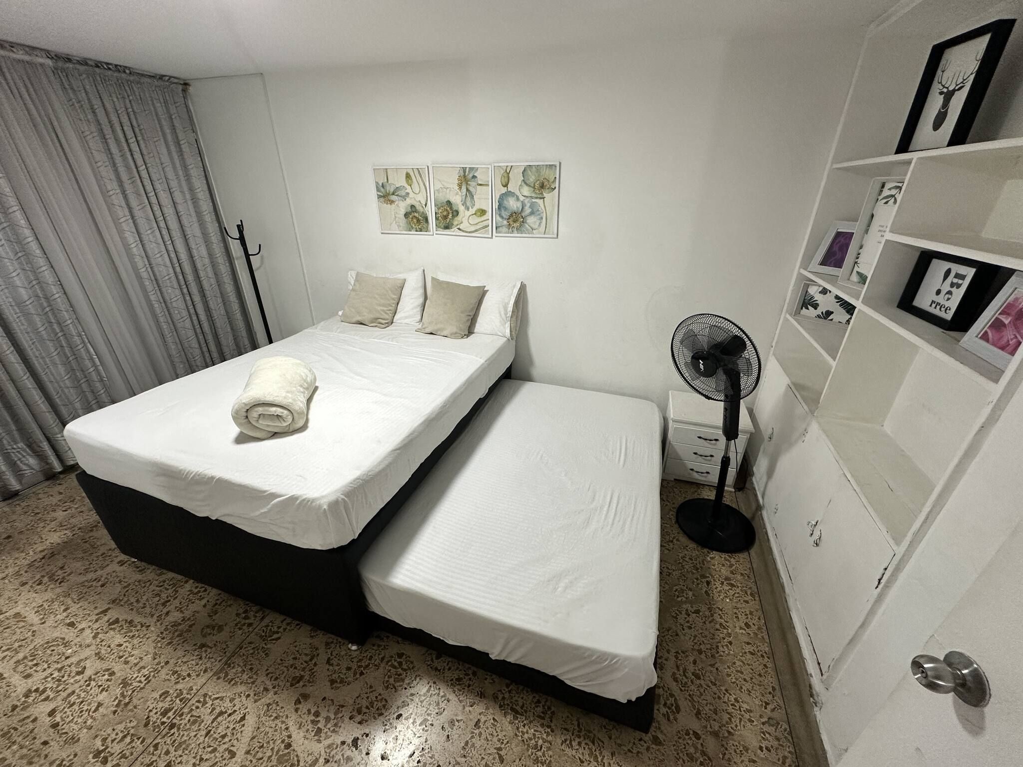 Ein Zimmer mit einem Bett, einem Nachttisch, einem Ventilator und einem Regal mit gerahmten Bildern.
