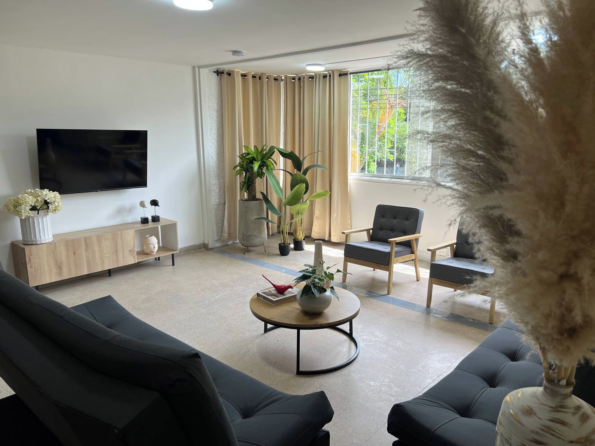 A modern living room with a flat-screen TV, a wooden console table, two black sofas, a round coffee table, and a large window with curtains.