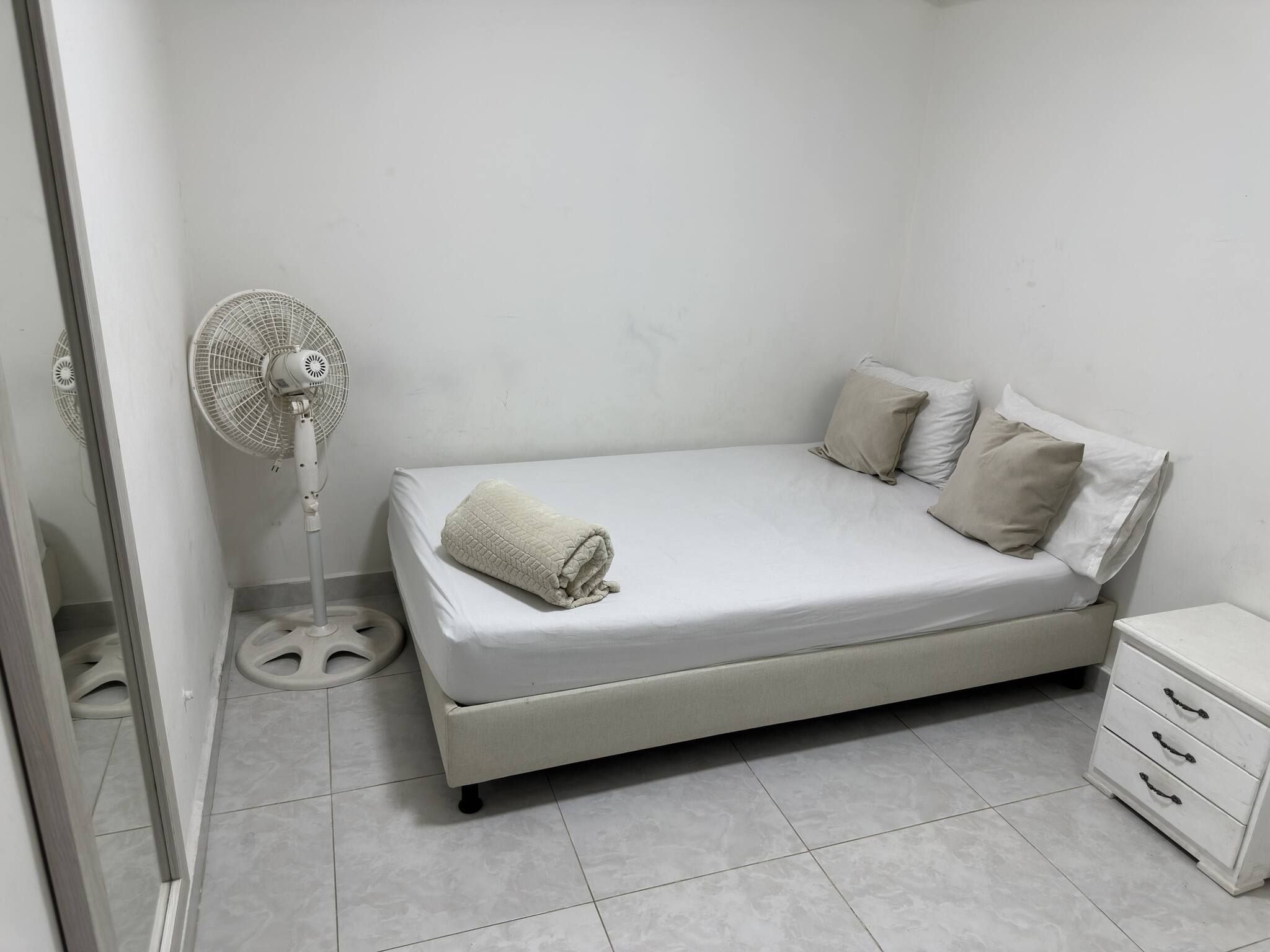 A small, neatly arranged bedroom with a single bed, a bedside table, and a standing fan.