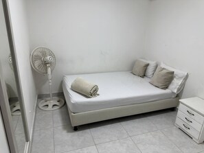 Un dormitorio pequeño y ordenado con una cama individual, una mesita de noche y un ventilador de pie.