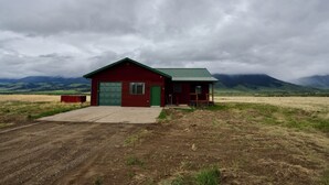 A red building with a green roof, a concrete driveway, and a grassy field in the foreground. - Newly Renovated 3-bedroom home near Yellowstone National Park (Livingston)