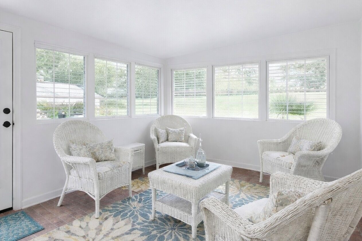 A room with white wicker furniture, large windows, and a patterned rug.