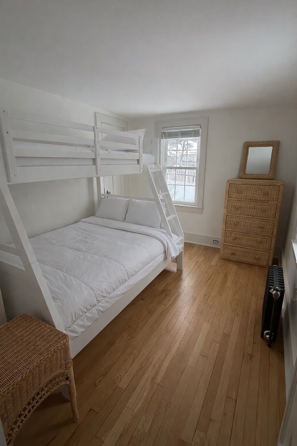 Ein Schlafzimmer mit einem Etagenbett, einem Korbstuhl, einer Kommode und einem Fenster mit Jalousien.