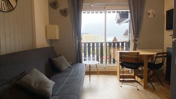 Una sala de estar acogedora con un sofá de color oscuro, una mesita redonda y un comedor con sillas. Un ventanal con cortinas deja entrar luz natural, y se ve un reloj de pared.