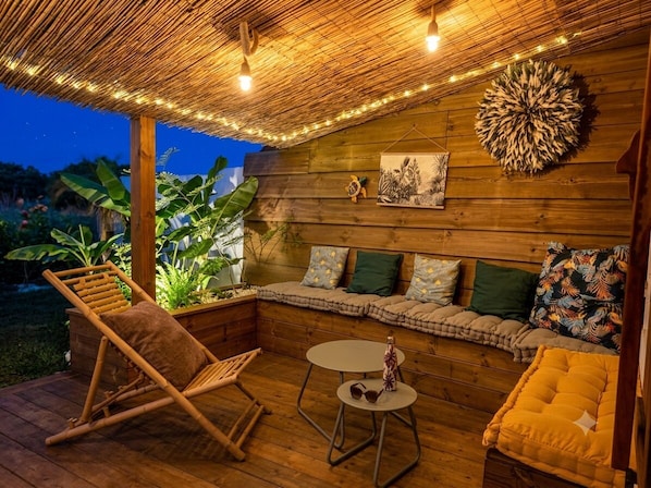 A wooden deck with a thatched roof, a cushioned seating area, and a small table with a bottle and sunglasses.