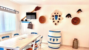 A dining area with a white table and blue chairs, a TV mounted on the wall, decorative wall hangings, and a large barrel. - Mamuthones close-up (Sas Linnas Siccas)