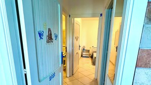 A hallway with a tiled floor, a white door with a vent, and a small table with a lamp. - Mamuthones close-up (Sas Linnas Siccas)