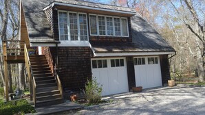 A two-story house with a wooden exterior, a front porch with steps, a garage, and a gravel driveway. - Adorable 1-bedroom cottage with AC in Shelter Island (Shelter Island)