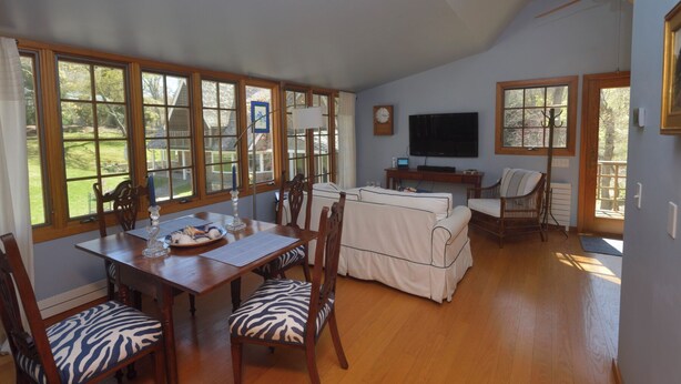 A spacious living room with a dining area, large windows, a flat-screen TV, and a ceiling fan. - Adorable 1-bedroom cottage with AC in Shelter Island (Shelter Island)