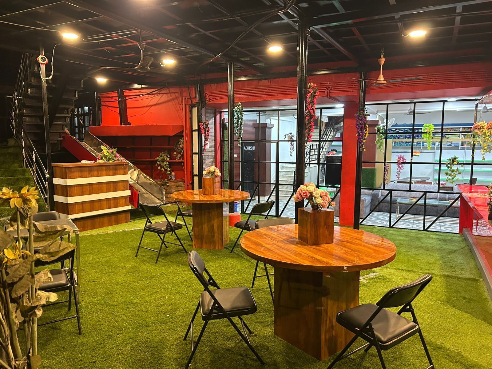 A modern indoor space with a green artificial grass floor, wooden tables, and black chairs. There are red accents on the walls and a staircase leading to an upper level.