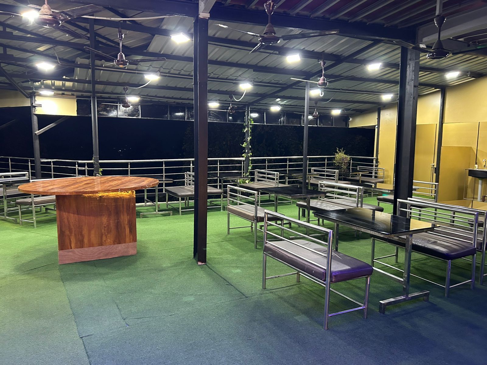 Un área de estar interior con mesas y bancos, una mesa de madera central y ventiladores de techo.