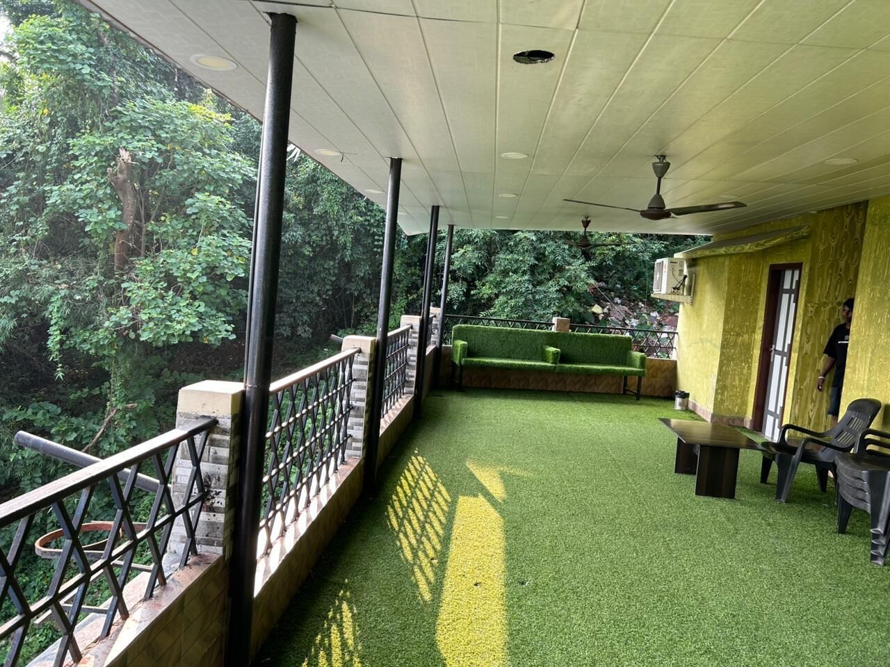 A covered outdoor area with artificial grass, a seating area with tables and chairs, and a view of greenery.