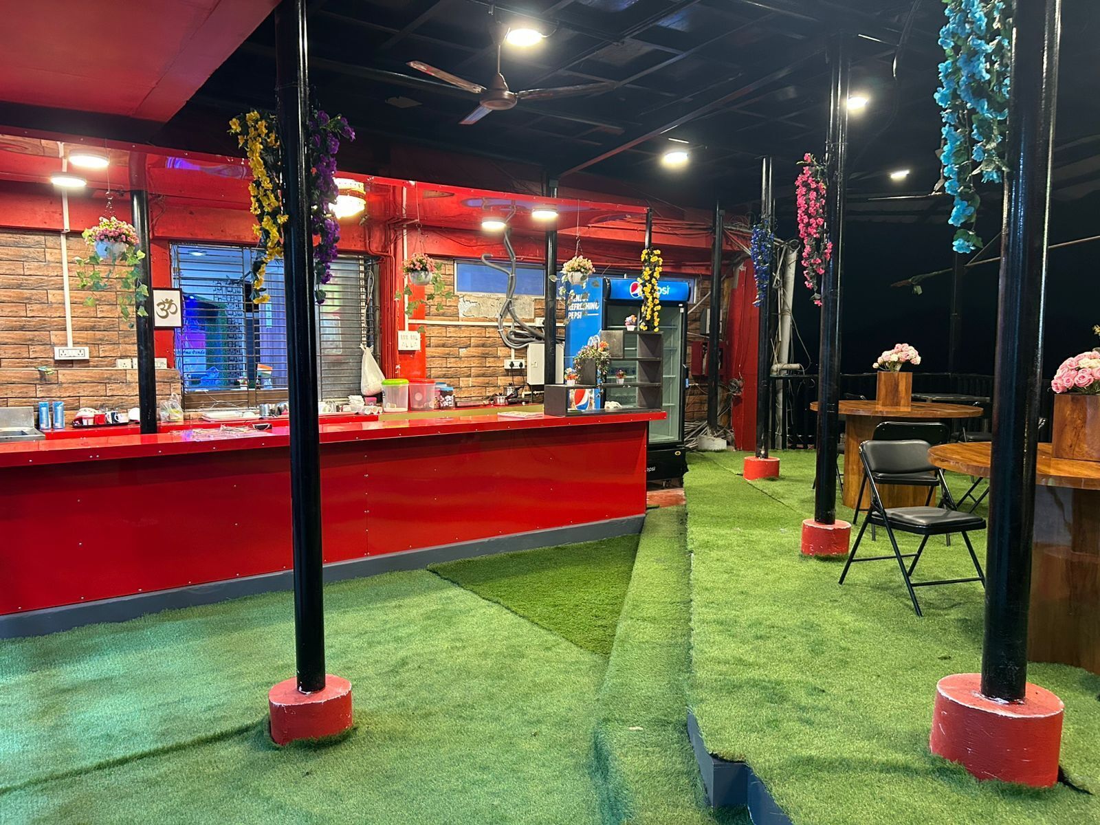 A restaurant interior with a red counter, green artificial grass flooring, and hanging flower arrangements.