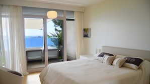 Un dormitorio con una cama grande, vista al océano a través de una puerta corrediza y una pequeña silla blanca.