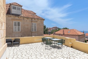 Un bâtiment en pierre avec un toit en tuiles, un espace extérieur aménagé avec des chaises et des tables, et une vue sur la mer.