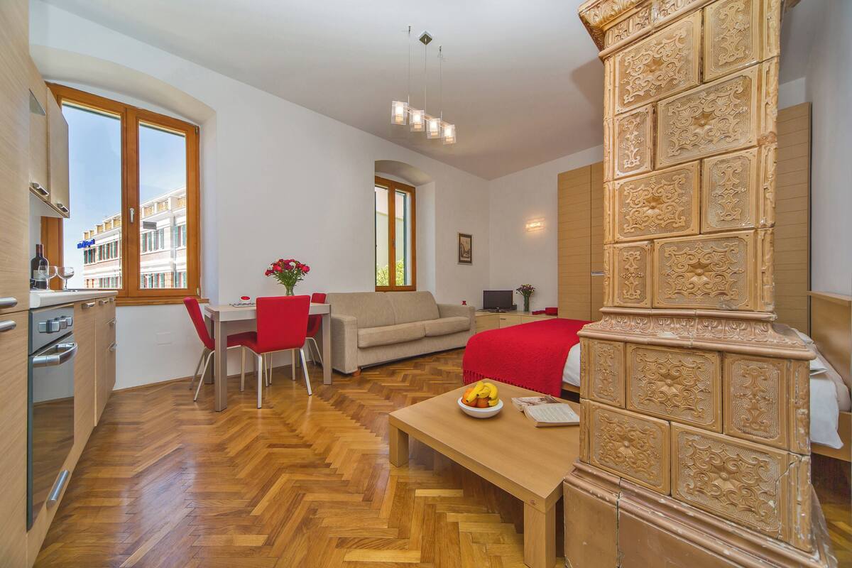 A modern hotel room with a wooden herringbone floor, a dining area with a red chair, a sofa, and a decorative column.