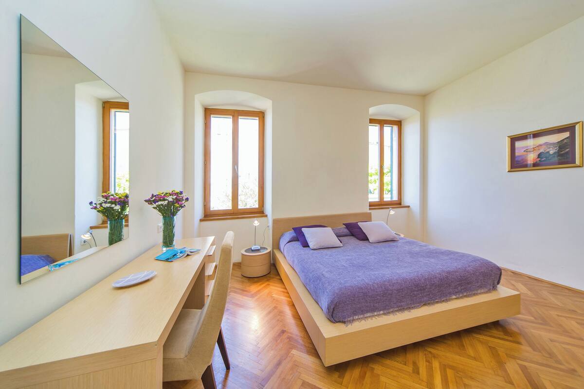A bedroom with a bed, a desk, a chair, a mirror, and two windows.