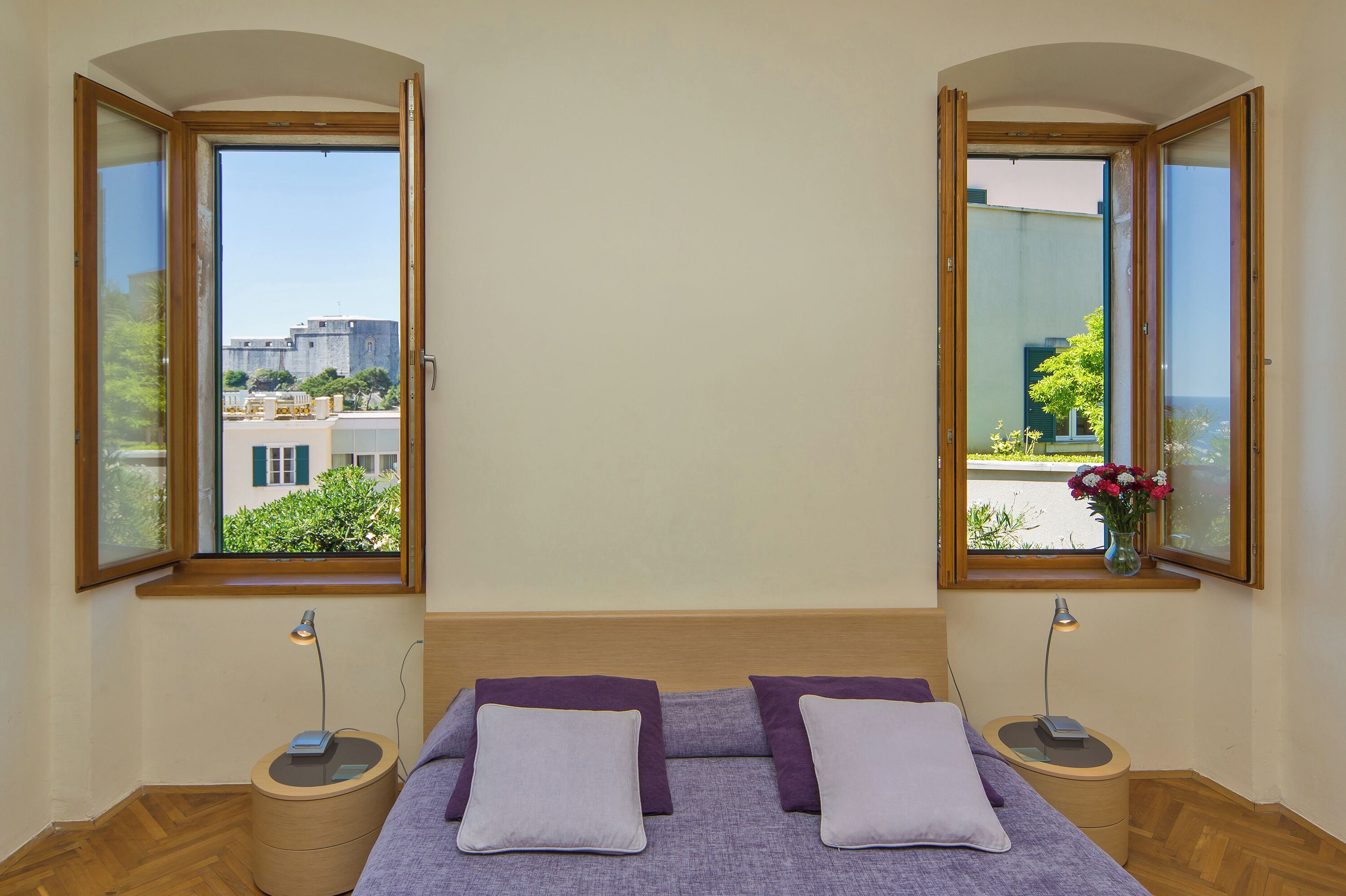 A bedroom with a bed, two nightstands, and two open windows with views of buildings and greenery.