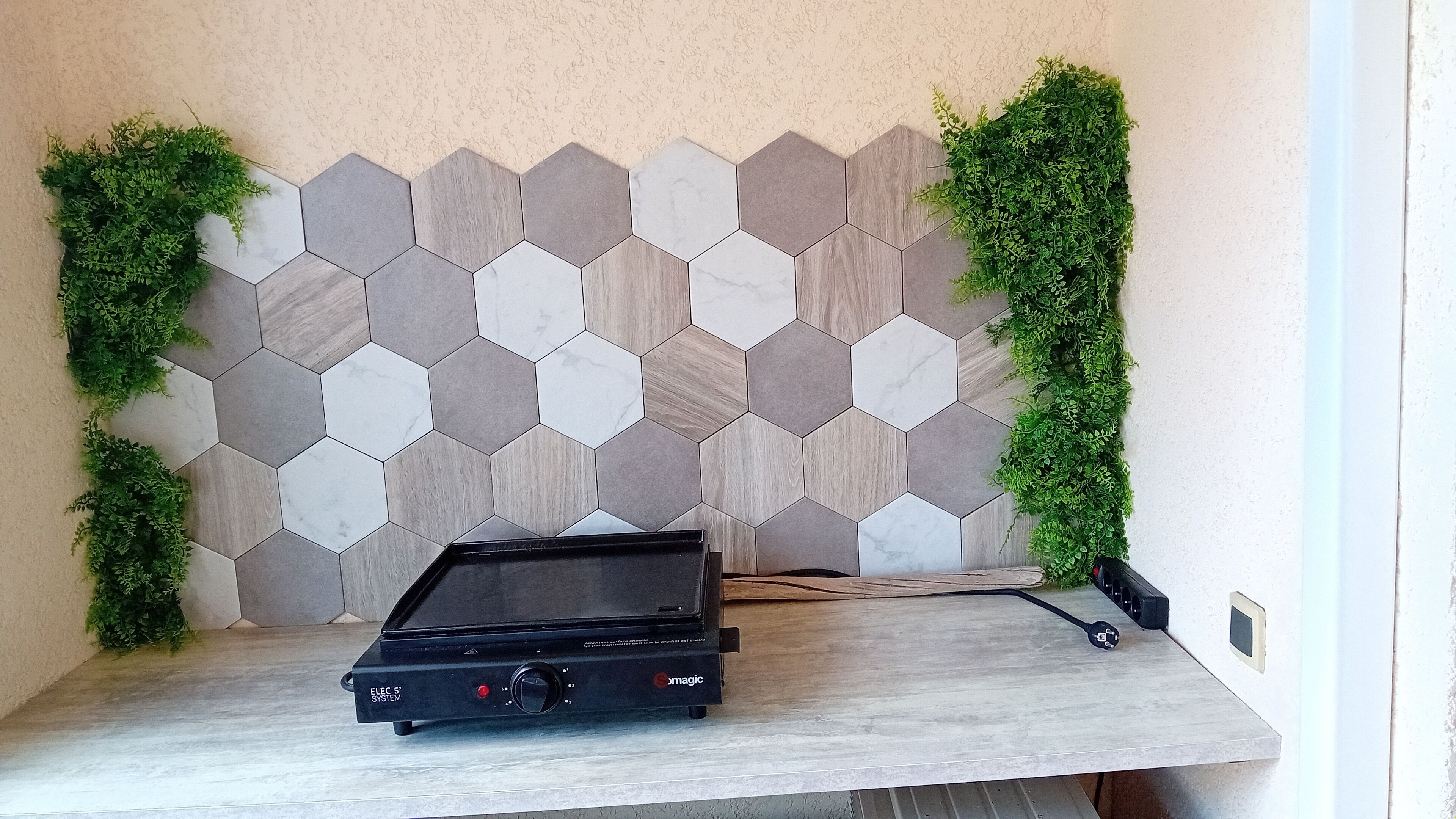 A room with a hexagonal tile wall, a black electronic device on a wooden table, and green plants on either side.
