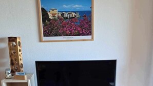 Un salon avec un téléviseur à écran plat posé sur un meuble blanc, un tableau encadré représentant Erbalunga, une petite étagère avec des objets décoratifs et un vase rempli de fleurs.