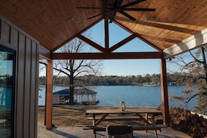 A covered outdoor area with a wooden ceiling, a table, and benches, overlooking a lake with a dock and a building in the background.