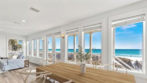 Un'ampia sala da pranzo con tavolo e sedie in legno, ampie finestre vista oceano e un'area salotto accogliente con divano e cuscini.
