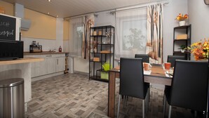 Cozinha moderna com piso de madeira, área de refeições com cadeiras pretas e uma estante preta com objetos decorativos.