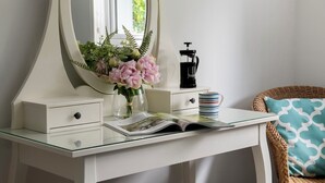 Une coiffeuse blanche avec un grand miroir, un petit vase avec des fleurs, une cafetière à piston et un magazine sur une table en verre.