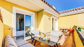 Un balcone luminoso con un divano, un tavolo e sedie, e una porta che dà su una stanza con vista.