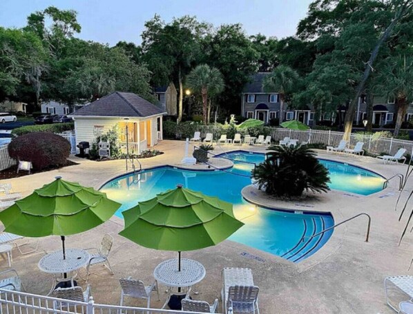 A swimming pool with two green umbrellas, lounge chairs, and a small building in the background.