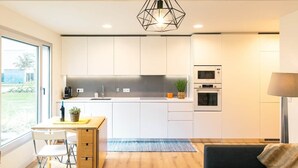 A modern kitchen with white cabinets, a wooden dining table, and a built-in oven. - Baia Rio by Proud Progress Homes (Seixal)