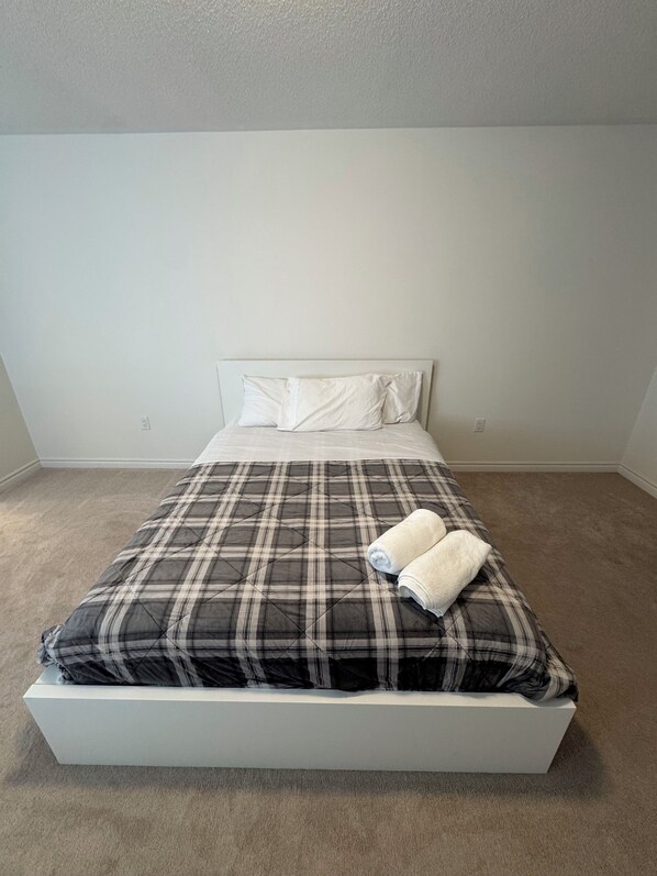 Una cama bien hecha con colcha a cuadros y dos toallas enrolladas, cabecera blanca y pared lisa con toma de corriente.