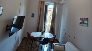 En kompakt opholdsstue med sofa, spisebord med stole, et fladskærms-tv på væggen, en loftlampe og et vindue med gardiner.