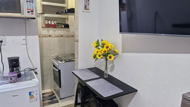 Een compacte keuken met een magnetron, koffiezetapparaat en een kleine koelkast. Er staat een eettafel met een vaas zonnebloemen en een stoel.