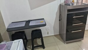Een kleine, netjes ingerichte kamer met een bureau, twee zwarte krukken, een aan de muur bevestigde televisie en een houten kast met lades.