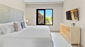 A modern bedroom with a large bed, a wooden dresser, a flat-screen TV, and a window with a view of a building and greenery. - Stylish Gold Coast Condo Sunrise by Bocobay (Noord)