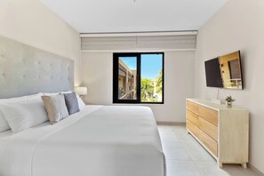 A modern bedroom with a large bed, a wooden dresser, a flat-screen TV, and a window with a view of a building and greenery. - Stylish Gold Coast Condo Sunrise by Bocobay (Noord)