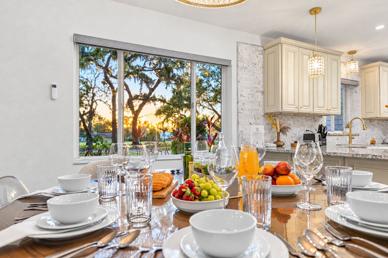 A dining area with a table set for a meal, a window with a view of trees, and kitchen cabinets in the background.