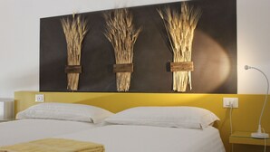 Une chambre d’hôtel avec un lit, des oreillers blancs, une tête de lit jaune et une œuvre d’art représentant des tiges de blé, accrochée au mur.
