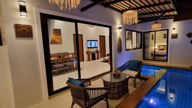 Ein moderner Innenbereich mit Schwimmbecken, einem Glastisch, Korbstühlen, einer Decke mit sichtbaren Balken und einem großen Fenster mit Blick auf den Pool.