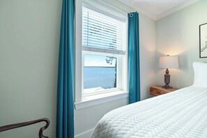 A bedroom with a bed, a nightstand with a lamp, a window with curtains, and a view of the sea. - Cozy Bayfront Guest Cottage - Boat Friendly Dock! (Elberta)